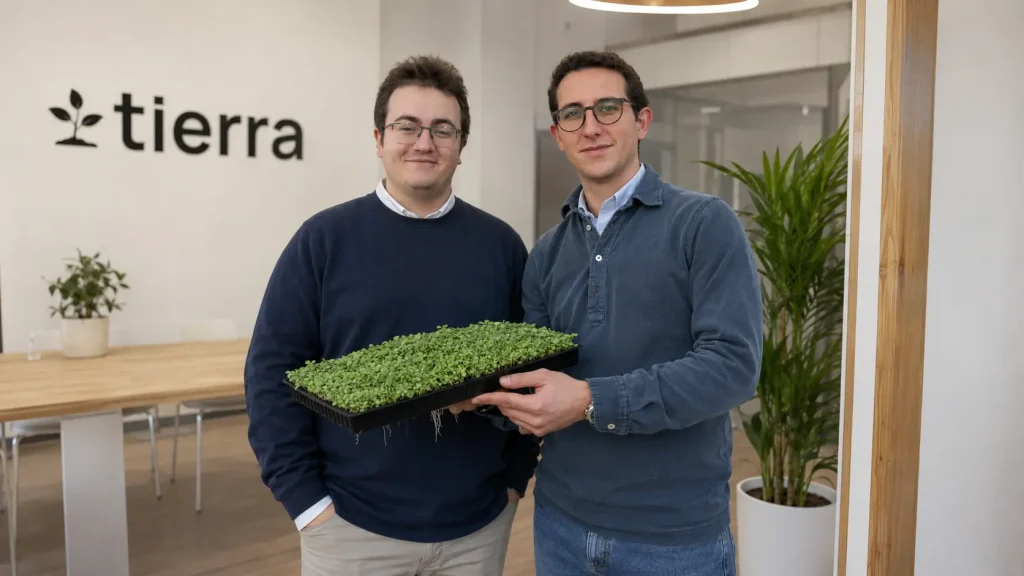 En una oficina luminosa, dos fundadores de Tierra sostienen una bandeja de microbrotes verdes mientras el logotipo se ve al fondo.